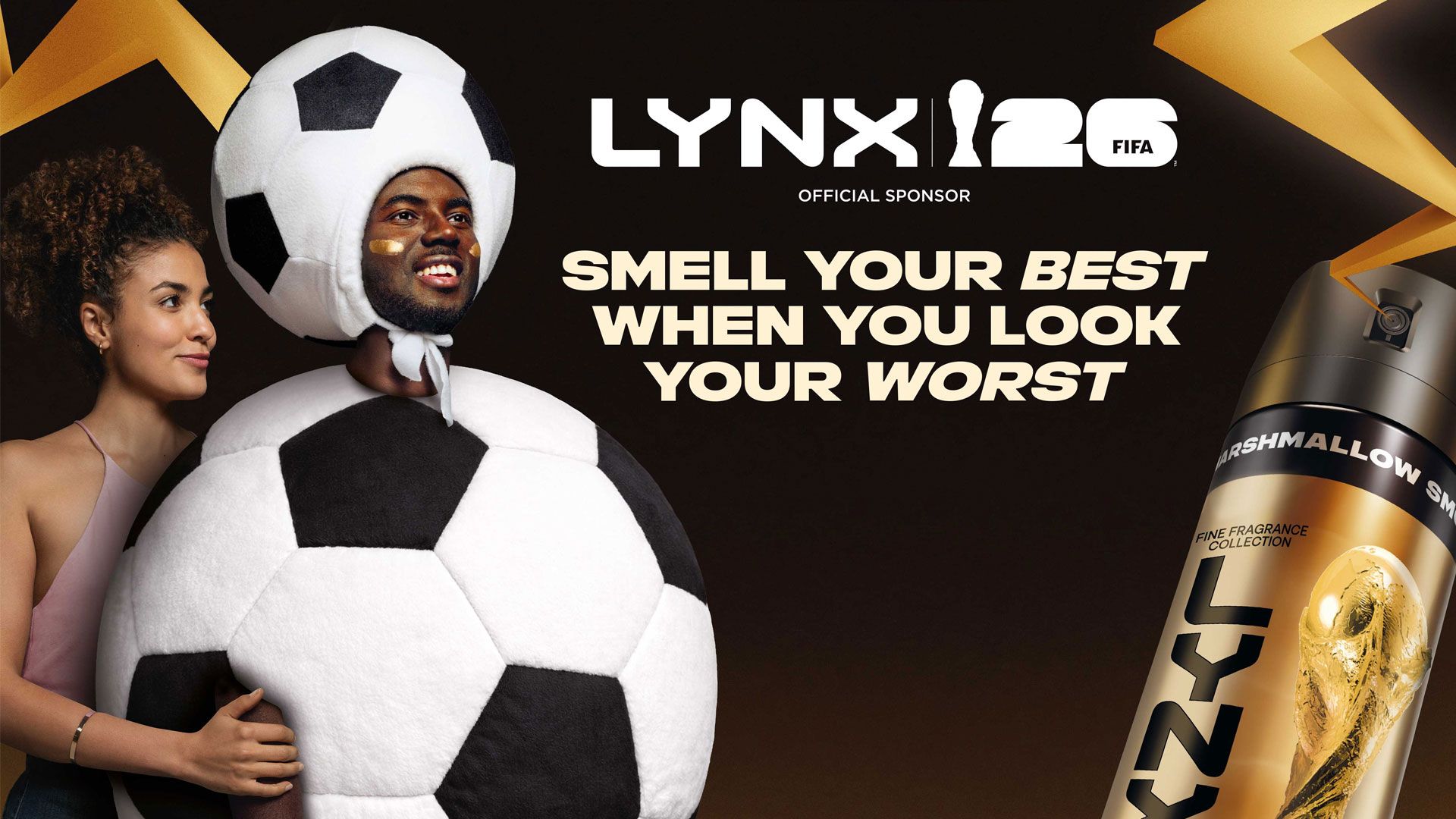 Two people stand side by side; one wears a large black and white football costume while the other holds their arm. On‑screen text reads “LYNX FIFA 26 Official Sponsor” and “Smell your best when you look your worst”, with a Lynx body spray can on the right.