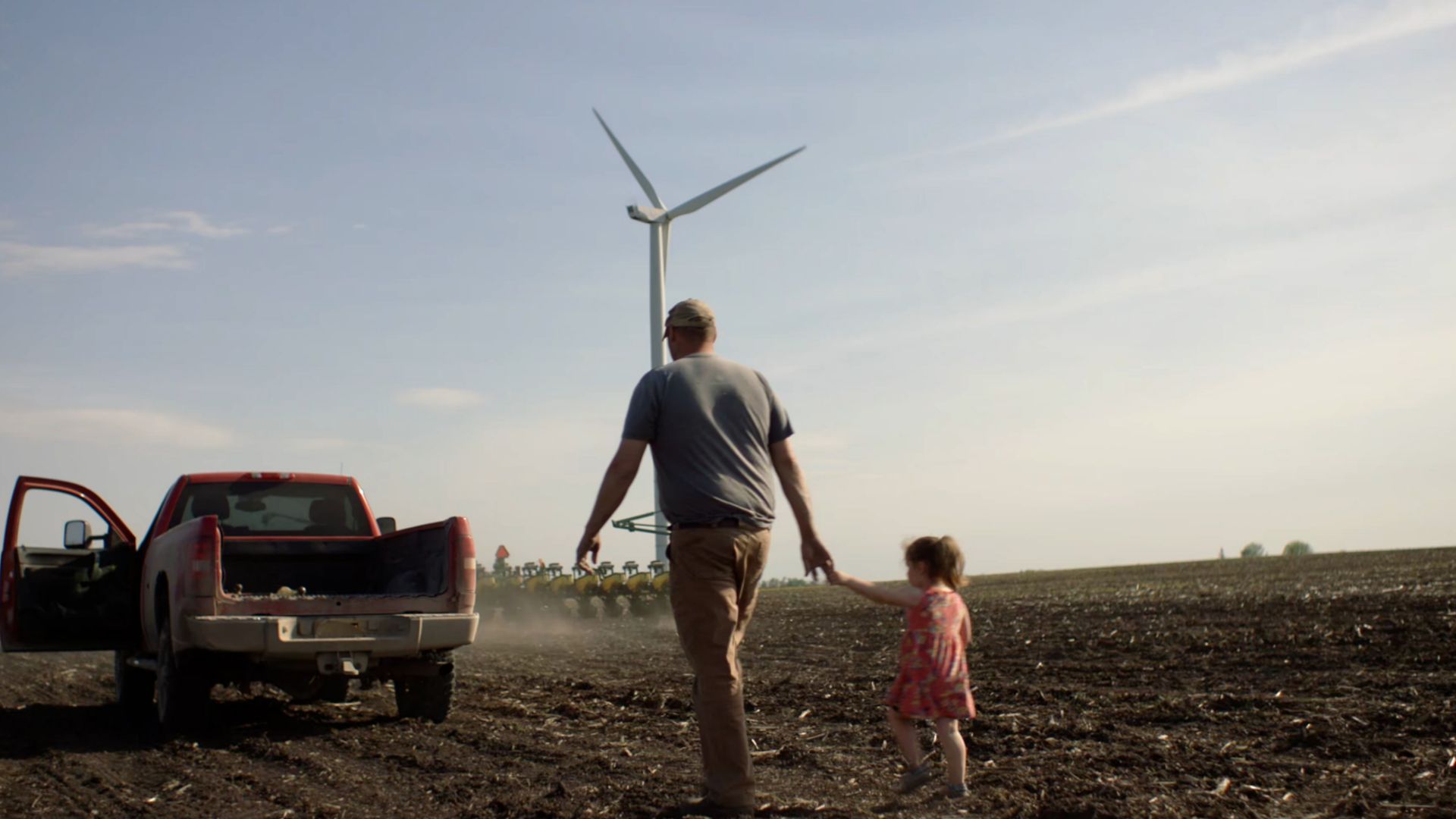 Father and daughter walking hand in hand across soil towards a red truck, wind turbine in the distance