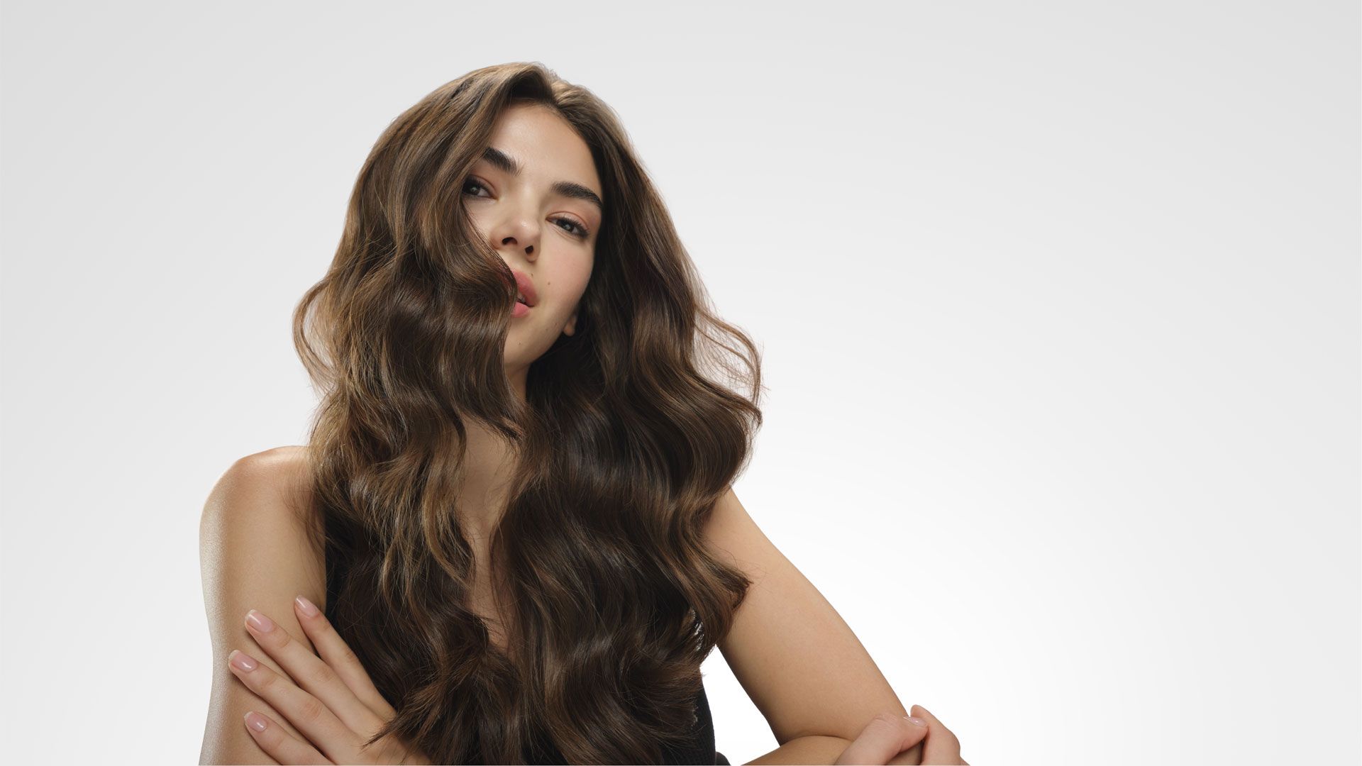 Person with long, glossy dark brown hair styled in loose waves, photographed against a light grey background with arms crossed to showcase hair length, shine and texture.