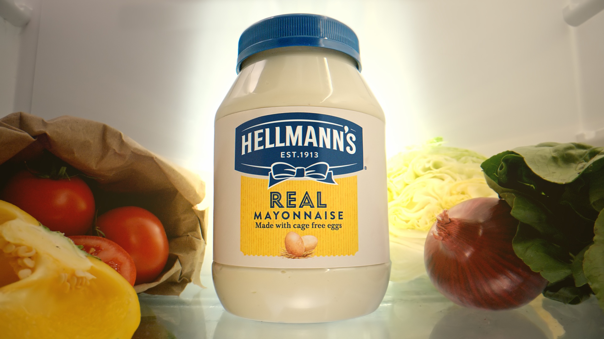 A bottle of Hellmann’s mayonnaise under a spotlight inside a fridge surrounded by vegetables including tomatoes, peppers, lettuce, cabbage and onion.