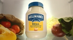 A bottle of Hellmann’s mayonnaise under a spotlight inside a fridge surrounded by vegetables including tomatoes, peppers, lettuce, cabbage and onion.