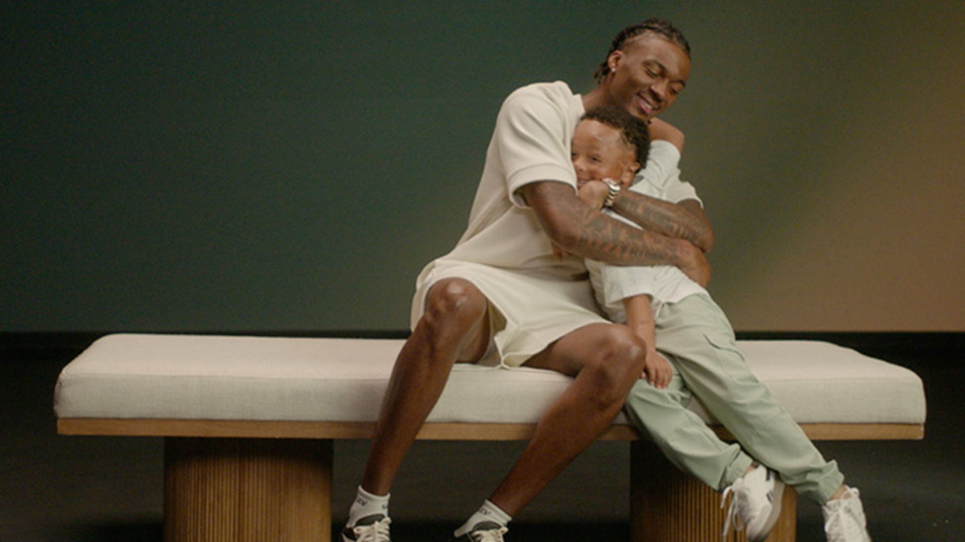 Two cousins hugging. One is much older than the other. They are both sitting on a white sofa.