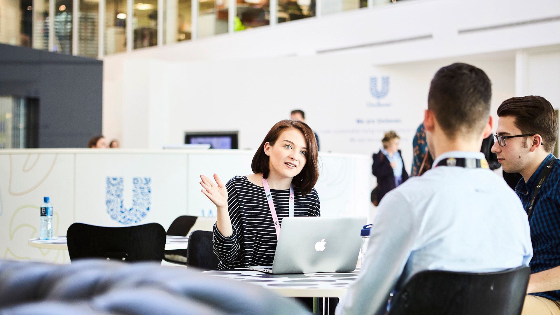 A group of people in a modern office engaged in discussion around a table, with one person gesturing while speaking. An open laptop is on the table, and the Unilever logo and branding is visible on a white wall in the background.