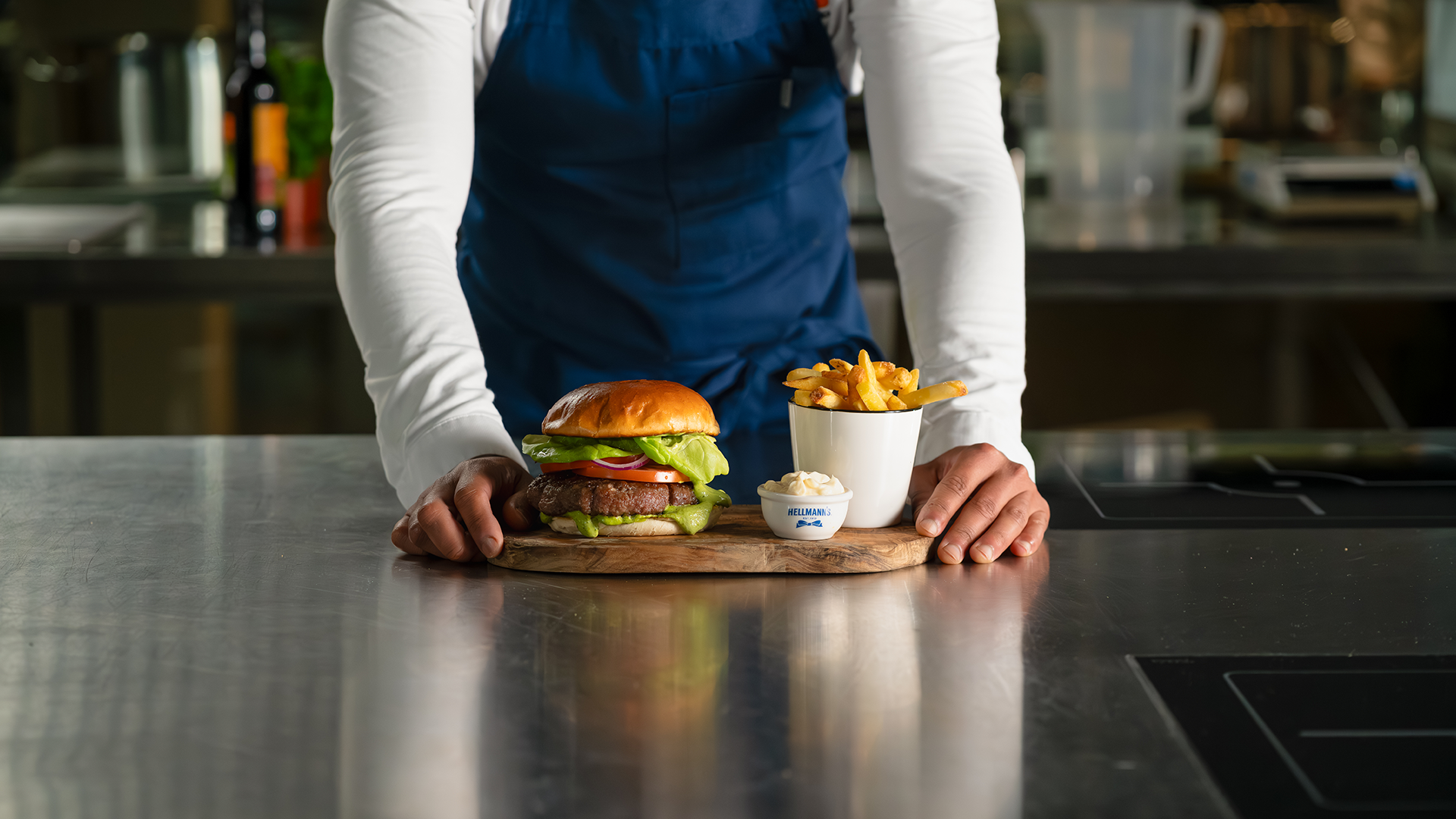 A person wearing a blue apron and leaning on a kitchen worktop. In front of them is a burger, a bowl of chips and a small serving of Hellmann’s mayonnaise, all arranged on a wooden chopping board.