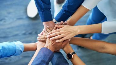 A group of people placing their hands in the centre of a circle in a pile. All are wearing clothes in shades of blue.