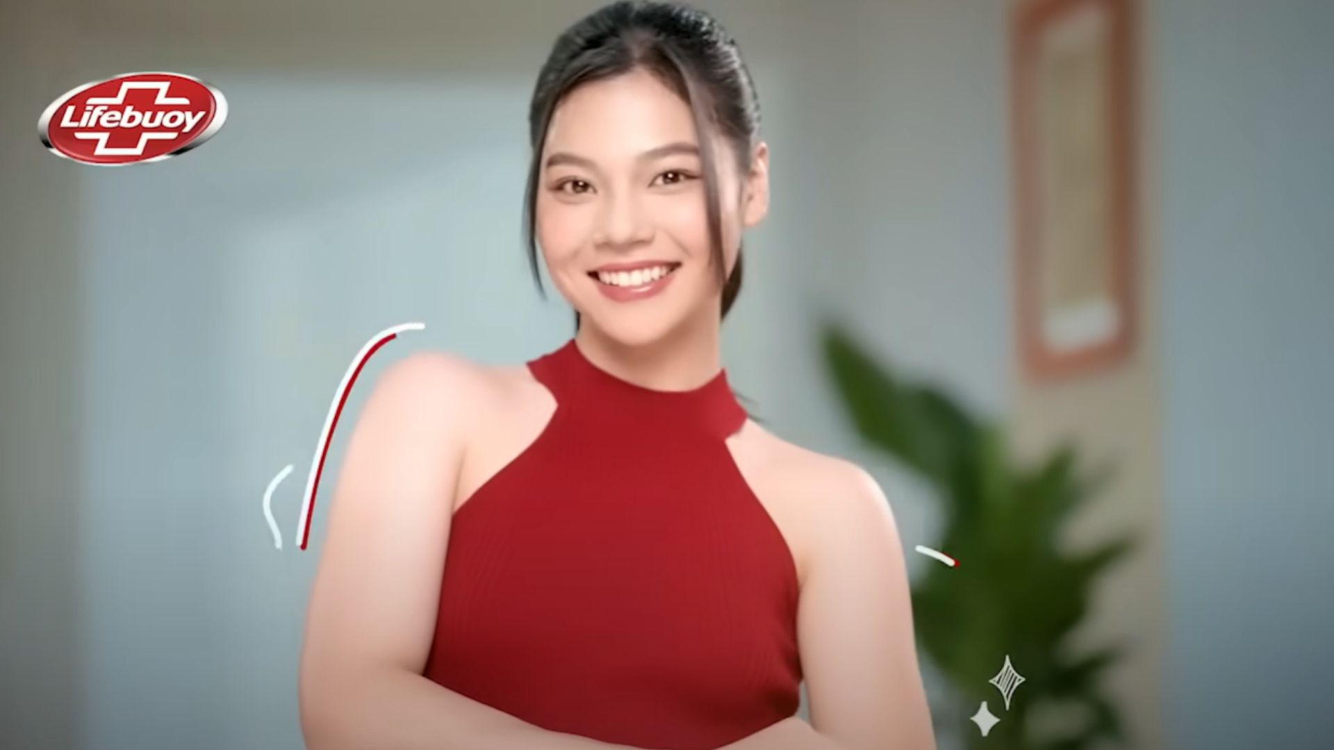 A lady smiling with her arms folded looking towards the camera. In the background is a picture hanging on the wall and a plant. There is a Lifebuoy logo in the top corner.