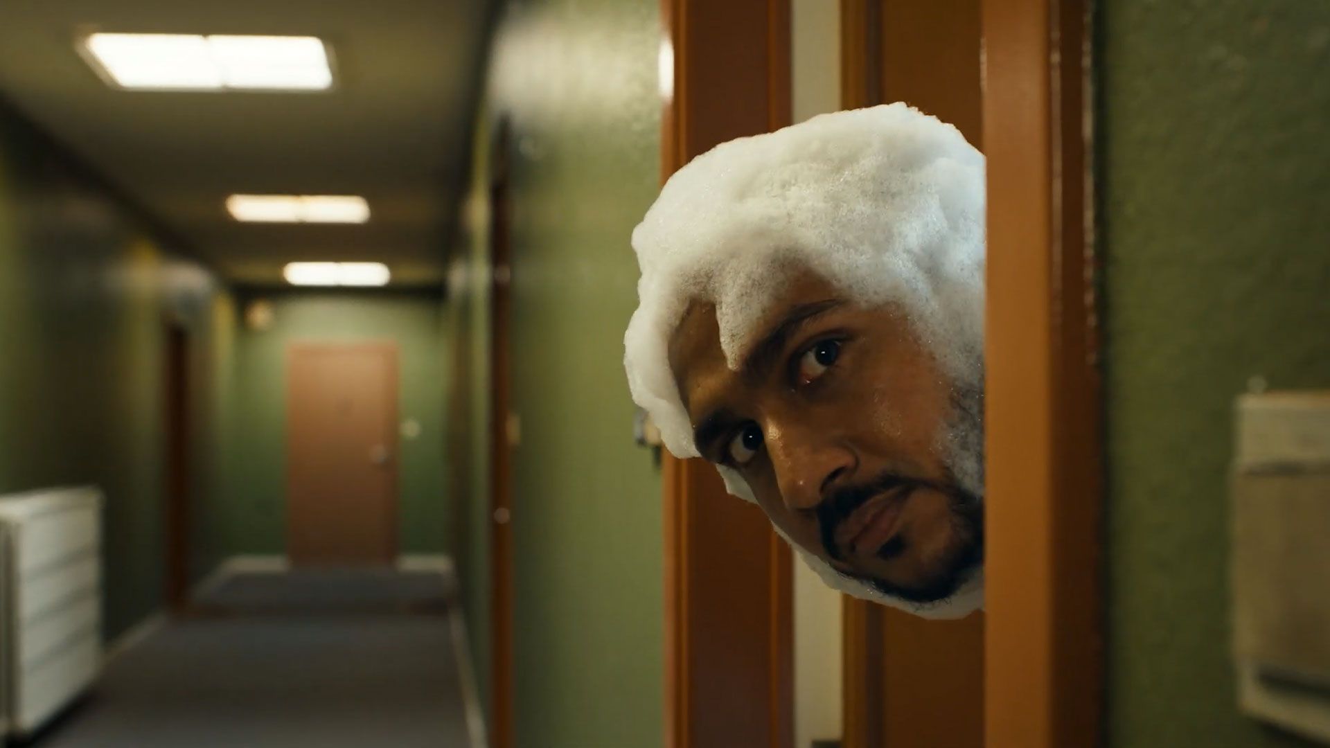 A person with a shaved beard and foam covering their head peeks through a slightly open doorway into a corridor with green walls and overhead lights.