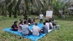 A group of people are seated in a circle on a blue tarpaulin sheet surrounded by palm trees. A facilitator is conducting a training session.