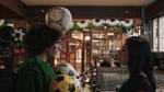 A person with a football balanced on their head and several footballs against their chest faces another person in a decorated pub with bunting and seated customers in the background.