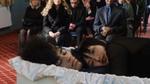 A funeral scene: a person lies in an open casket while a second person lies beside them inside the casket, looking toward the camera. Mourners in dark clothing sit in rows of chairs in the background.