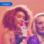 Hand holding a K18 hair product spray bottle with mist visible, set against a warm pink background. The image is tagged ‘Category’.