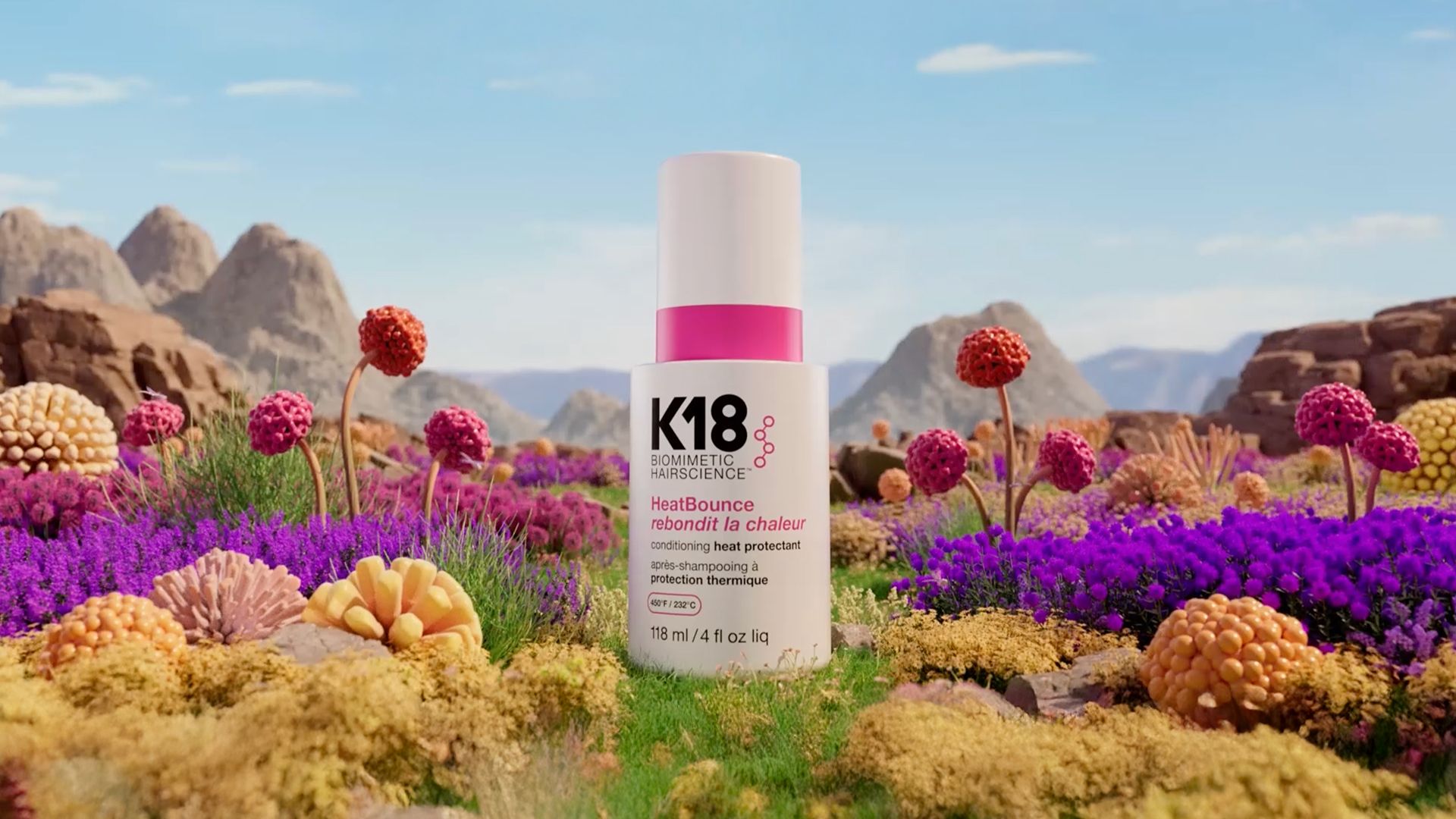 Bottle of K18 HeatBounce conditioning heat protectant displayed in a vibrant, colourful landscape with rocky mountains in the background and clusters of bright, spherical plants in shades of purple, pink, and yellow in the foreground.