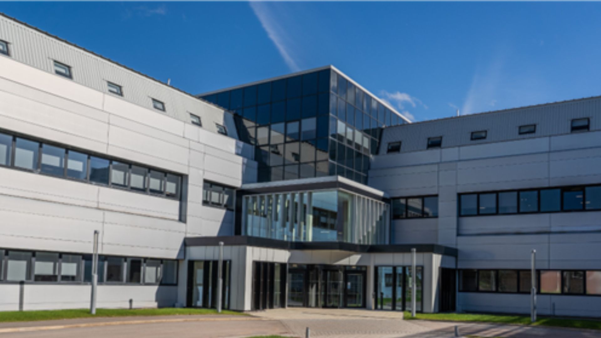 A low-level office building with a central main entrance, built predominantly from glass, with two grey wings extending to the left and right.