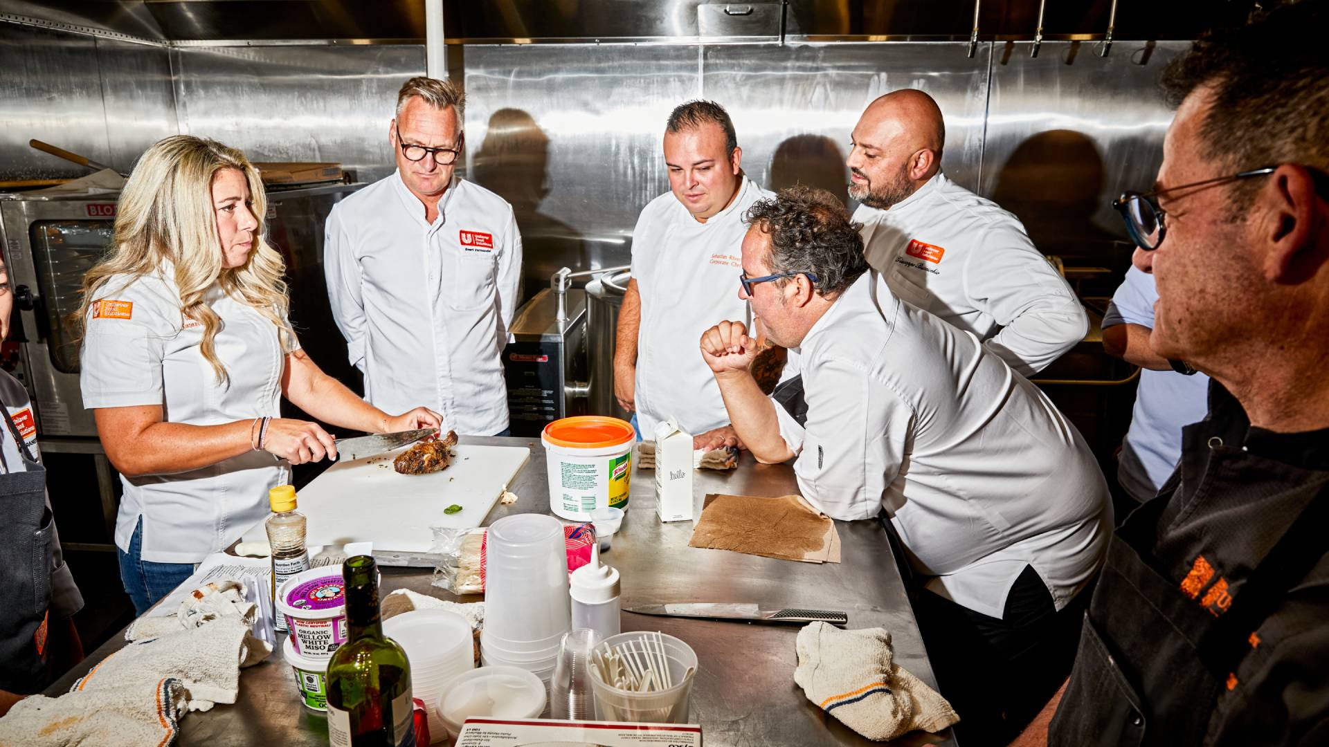 A group of Unilever Food Solutions chefs are gathered around a stainless-steel table as a colleague prepares a chicken dish. 