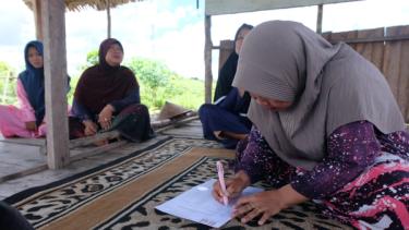 A photograph of four women in head coverings sitting cross-legged on a rug under a raised shelter, with trees in the background. The woman in the foreground is writing on paper.