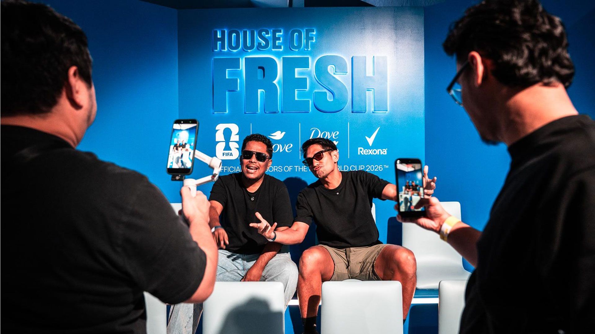 Two people sit on a white seating area in front of a blue “House of Fresh” event backdrop featuring FIFA, Dove and Rexona logos, while attendees record them on smartphones. The backdrop includes the text “Official Sponsors of the FIFA World Cup 2026™”.