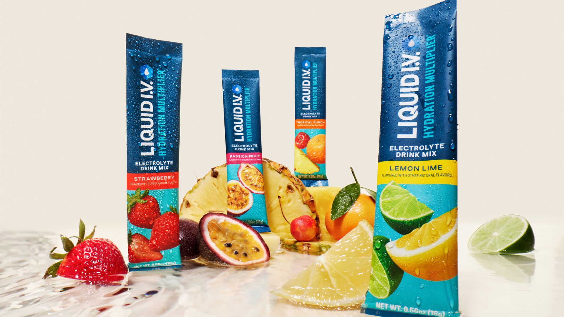 A variety of Liquid I.V. Hydration Multiplier electrolyte sachets in different flavours, including Strawberry and Lemon Lime. The packets are arranged around fresh fruits like strawberries, passion fruit, pineapple, lemon, lime, and orange slices on a light-coloured surface with water droplets.