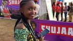 Young woman smiling at the camera, holding a mobile phone, in front of a Shujazz banner at an outdoor event.
