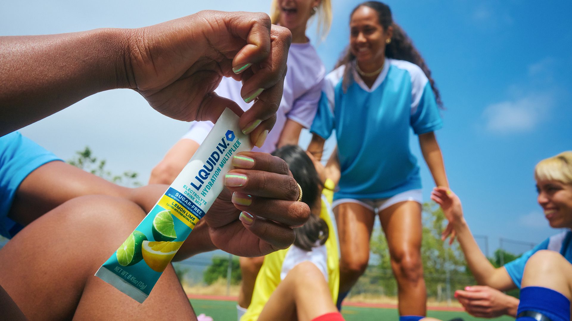 Close-up of a hand holding a Liquid I.V. Hydration Multiplier packet in lemon-lime flavour, with a group of people in sports uniforms gathered in the background on an outdoor field under a clear blue sky.