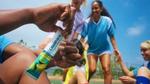 Close-up of a hand holding a Liquid I.V. Hydration Multiplier packet in lemon-lime flavour, with a group of people in sports uniforms gathered in the background on an outdoor field under a clear blue sky.