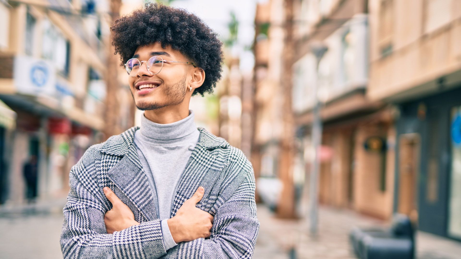 A young man with curly hair and glasses smiles confidently while standing on a city street with his arms crossed