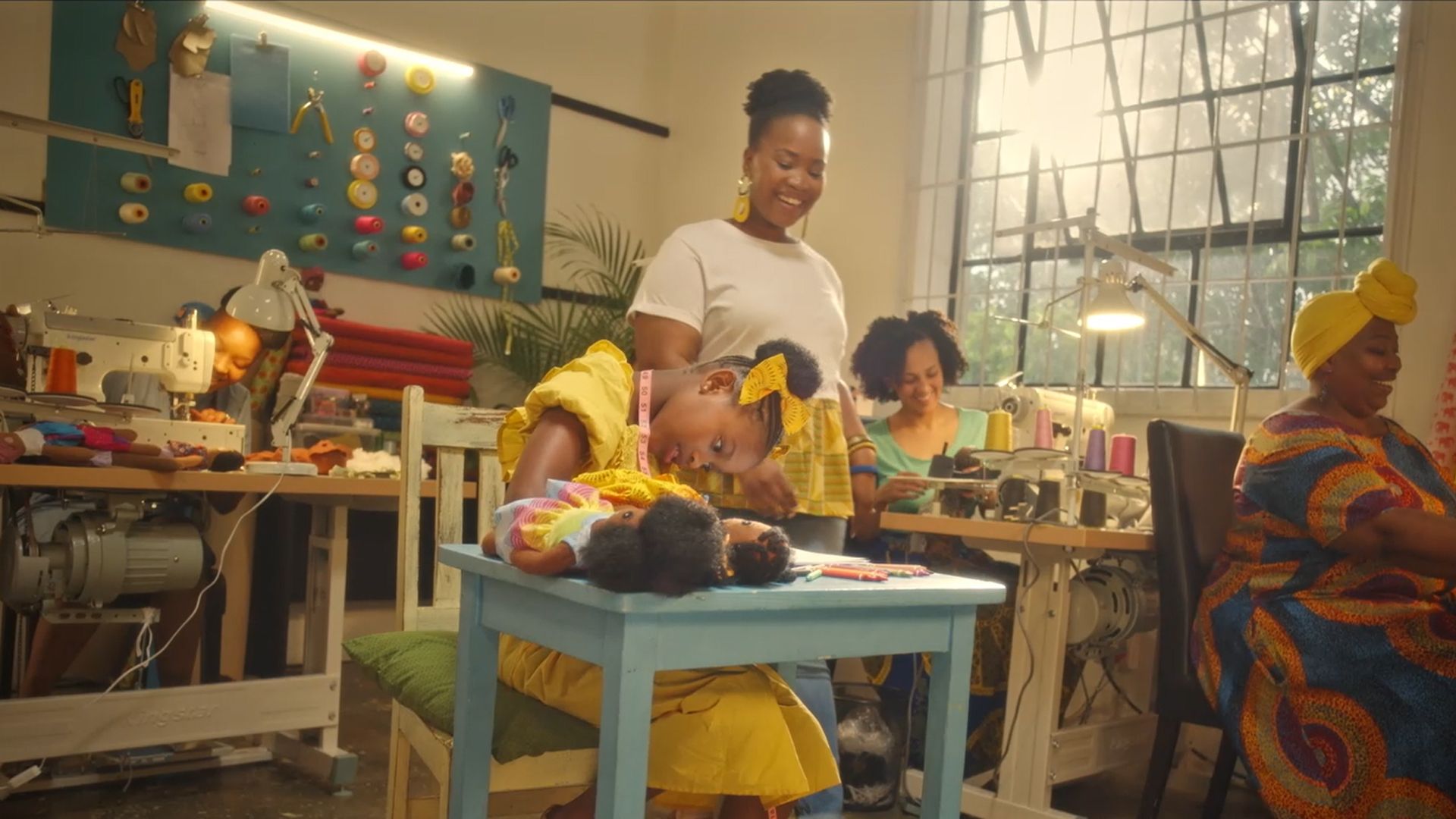 Three women working on sewing machines smile at a child learning to sew by hand