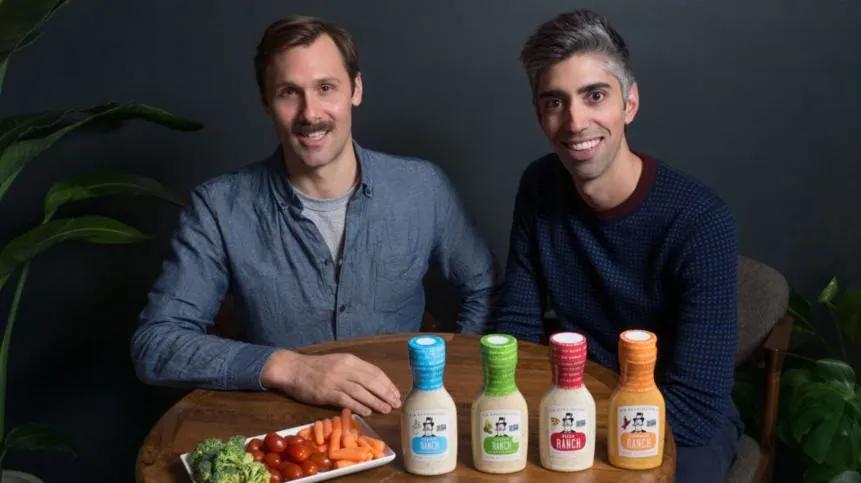 Two men with a collection of mayo bottles smiling