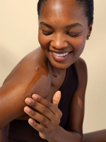A woman smiles as she rubs cream into her bare shoulder.