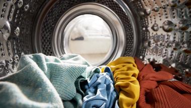 Close-up view from inside a washing machine drum, showing laundry in shades of green, blue, yellow and red ready to be washed.
