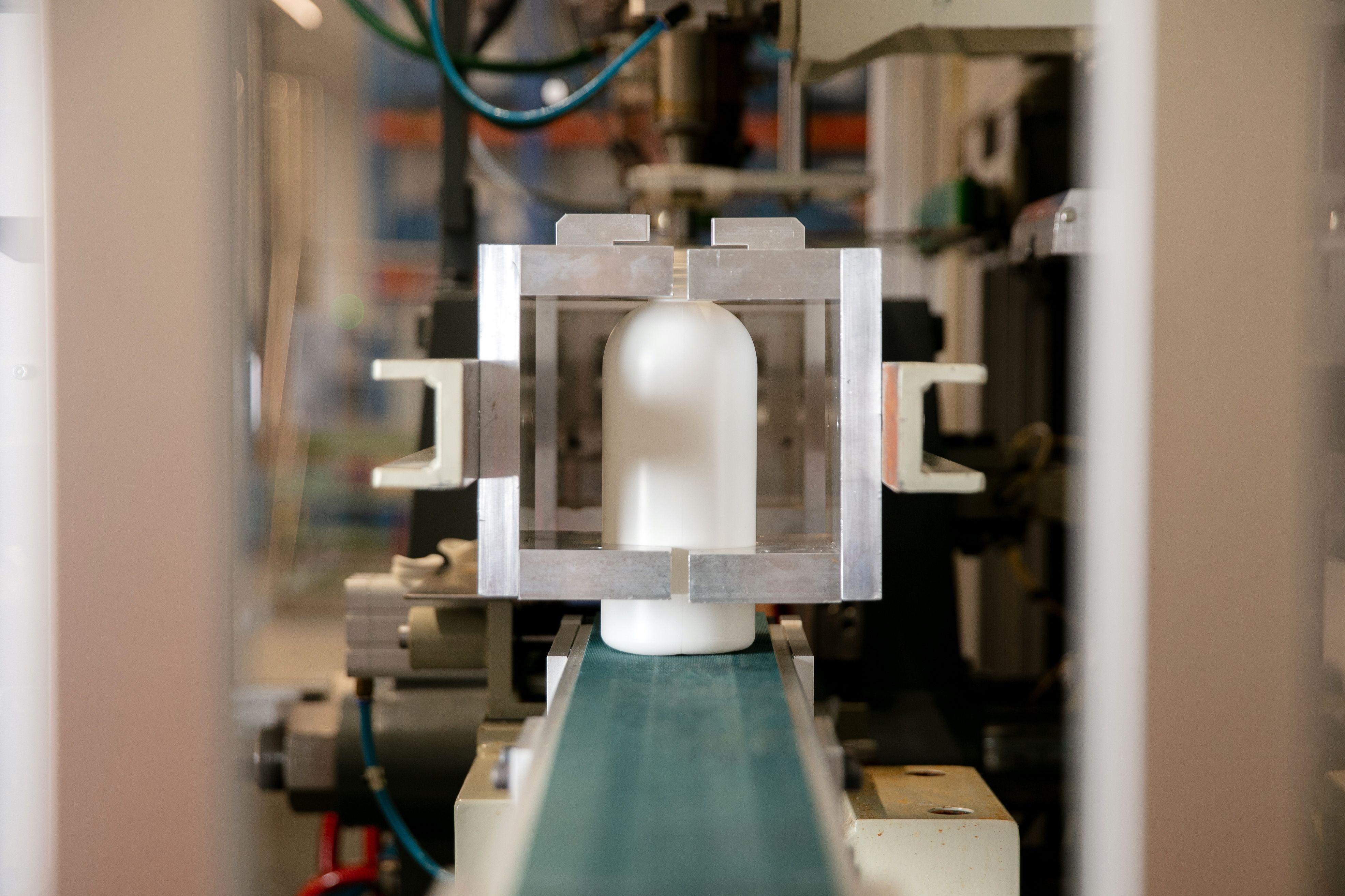 A close-up image of a white plastic bottle in an industrial testing setting. The bottle is held in place by a rectangular metallic frame.