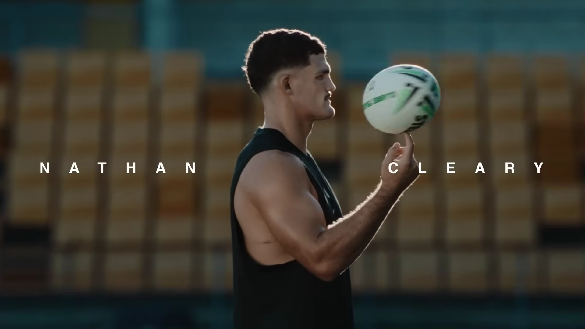 In the foreground, Nathan Cleary is spinning a rugby ball on his finger, with His name written on either side of him. In the background are rows of yellow stadium chairs.
