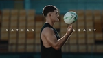 In the foreground, Nathan Cleary is spinning a rugby ball on his finger, with His name written on either side of him. In the background are rows of yellow stadium chairs.
