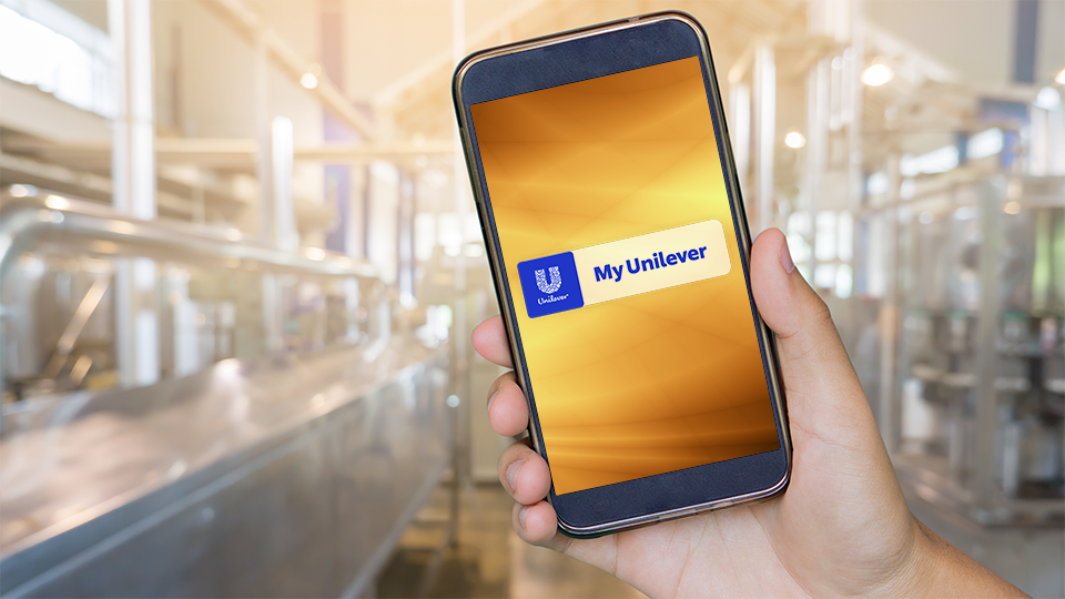 A hand holding an iPhone with a yellow screen with the Unilever logo and words ‘My Unilever’ against a blurred factory background. 