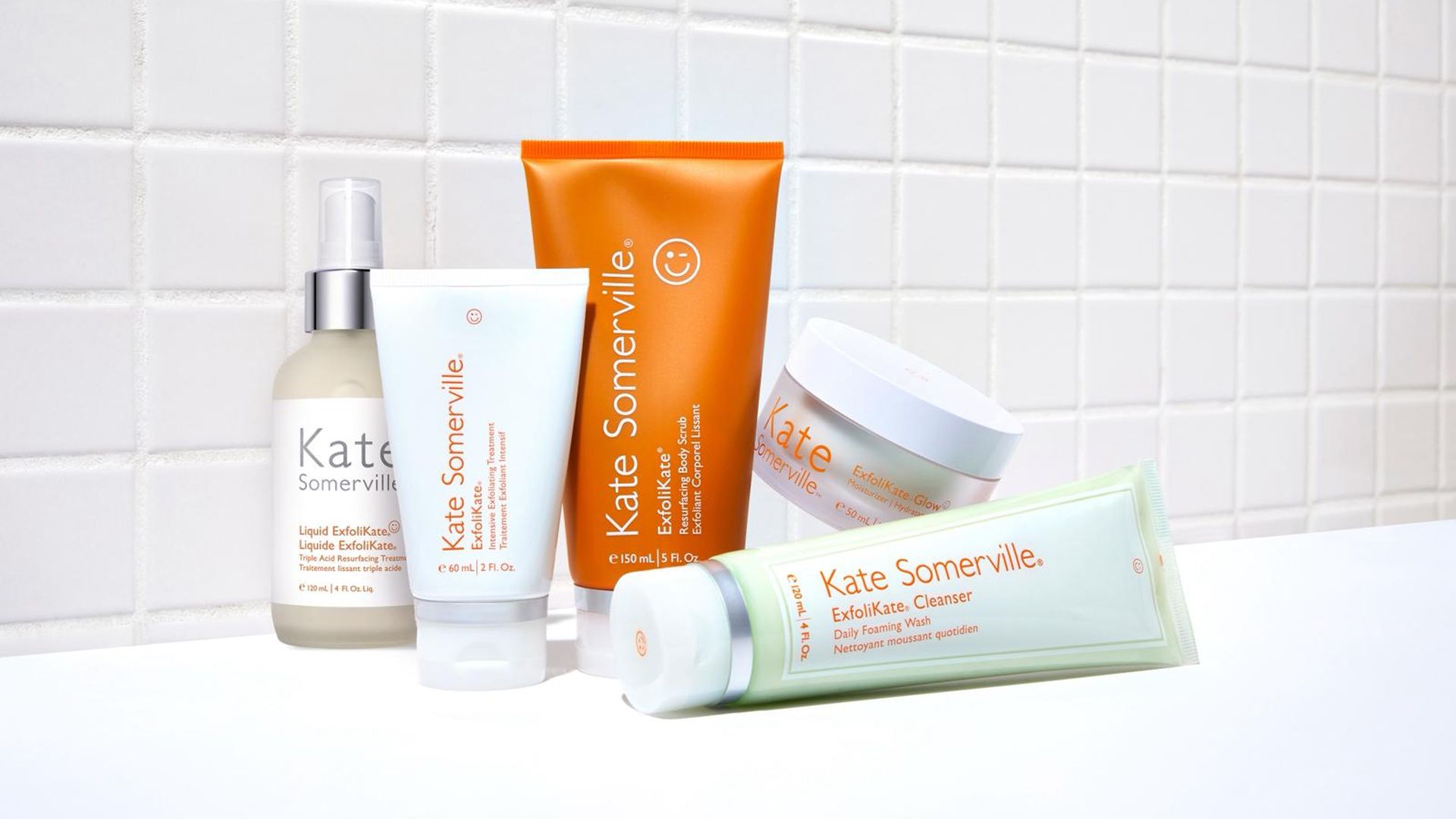 A selection of Kate Somerville skincare products arranged on a white surface against a tiled background. Items include a pump bottle of Liquid ExfoliKate, a white tube of ExfoliKate Intensive Exfoliating Treatment, an orange tube of ExfoliKate Glow Moisturiser, a white jar of Exfolikate Glow, and a green-and-white tube of ExfoliKate Cleanser.