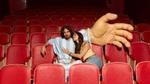 Two people sit together in a row of red cinema seats, one wearing a blue‑and‑white striped football‑style outfit, beside a large oversized hand prop resting across nearby seats.