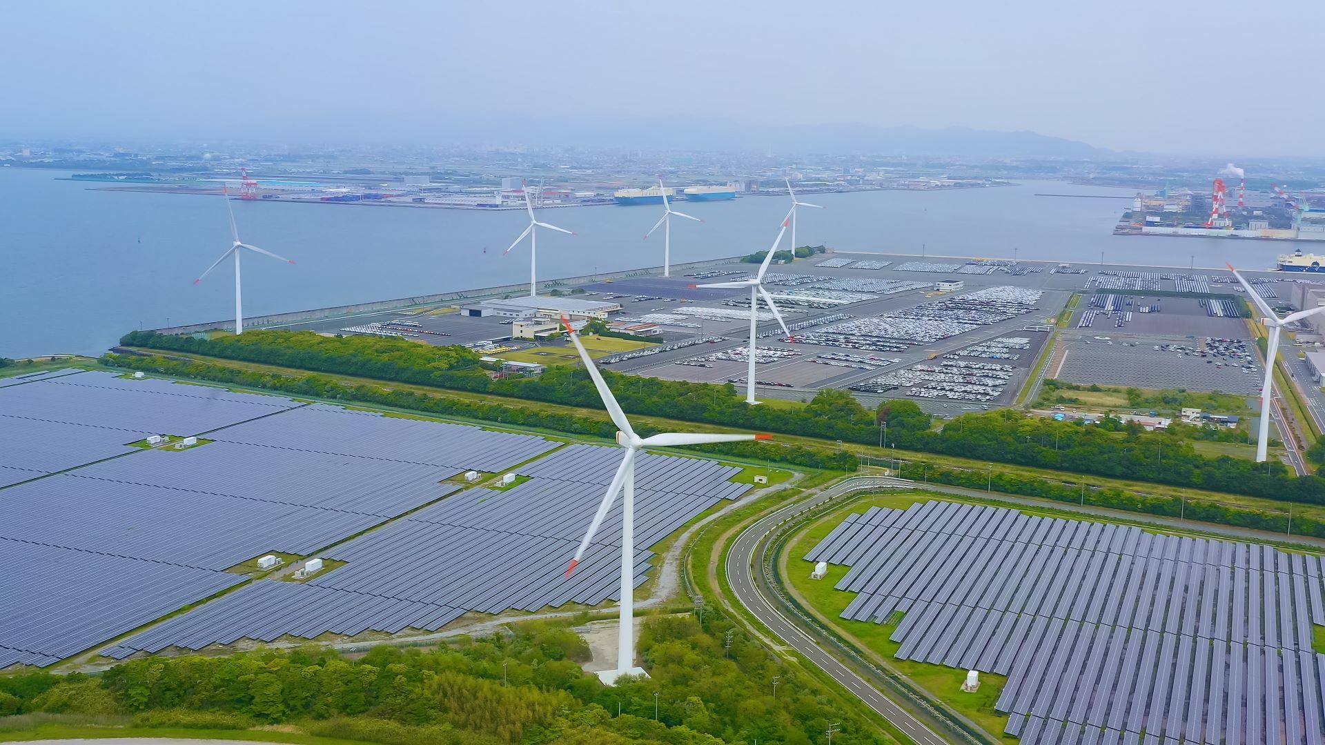 An aerial view of a renewable energy site featuring rows of solar panels in the foreground and multiple wind turbines in the background near a body of water, with an industrial port visible in the distance.