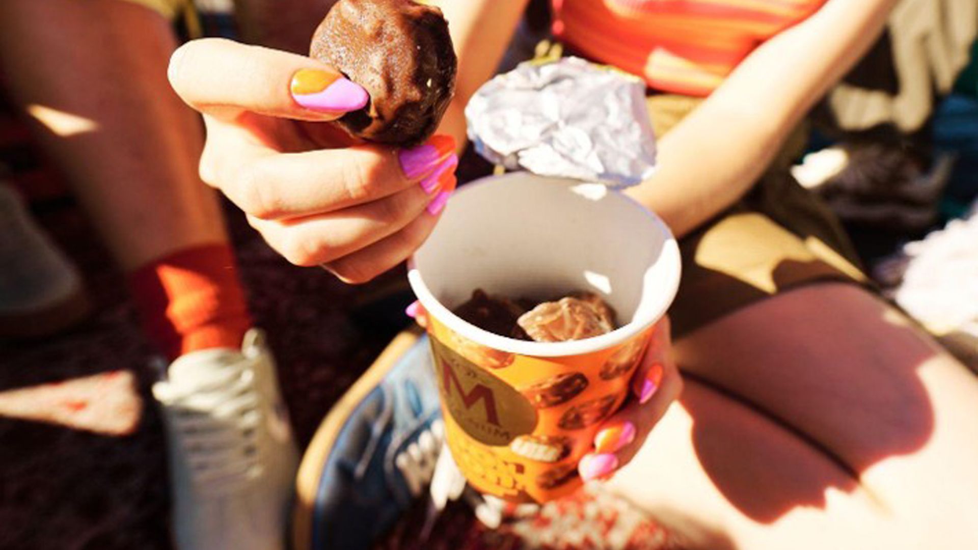 Person with brightly painted nails holding a chocolate-covered Magnum Bon Bon, seated on the ground in shorts and trainers under bright sunlight.