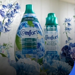 Comfort fabric conditioner and scent booster bottles surrounded by blue flowers, presented in a fresh, bright setting.