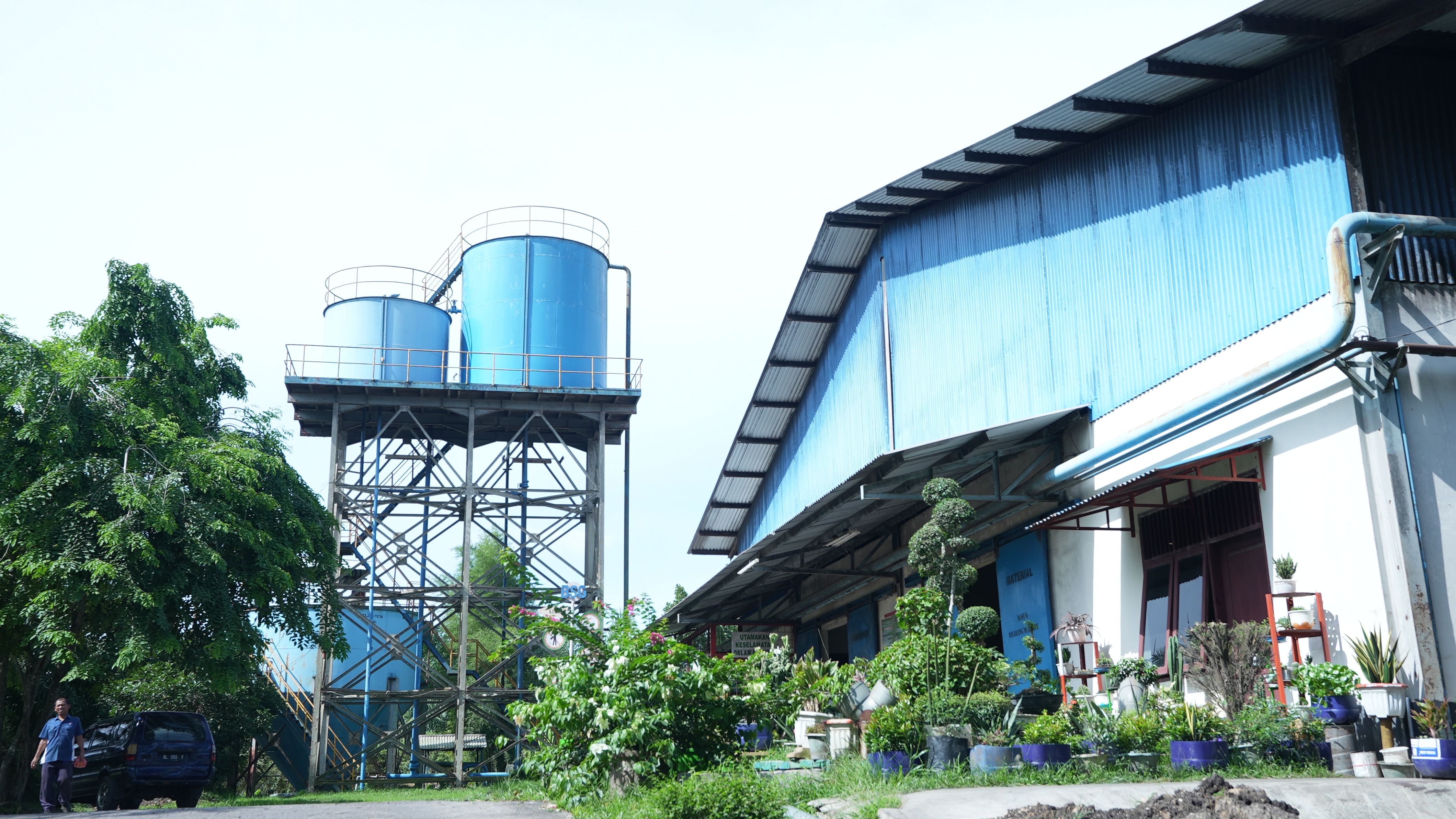 A photograph of industrial blue buildings at a mill in Aceh, Indonesia, with vegetation in the foreground.