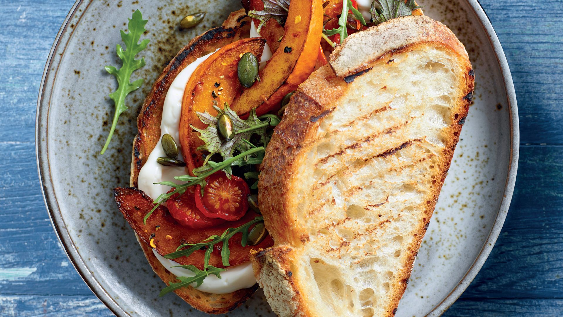 A delicious-looking open sandwich on a ceramic plate, featuring grilled bread topped with roasted pumpkin slices, cherry tomatoes, rocket, and pumpkin seeds. The vibrant colours and textures make it an appetising dish.