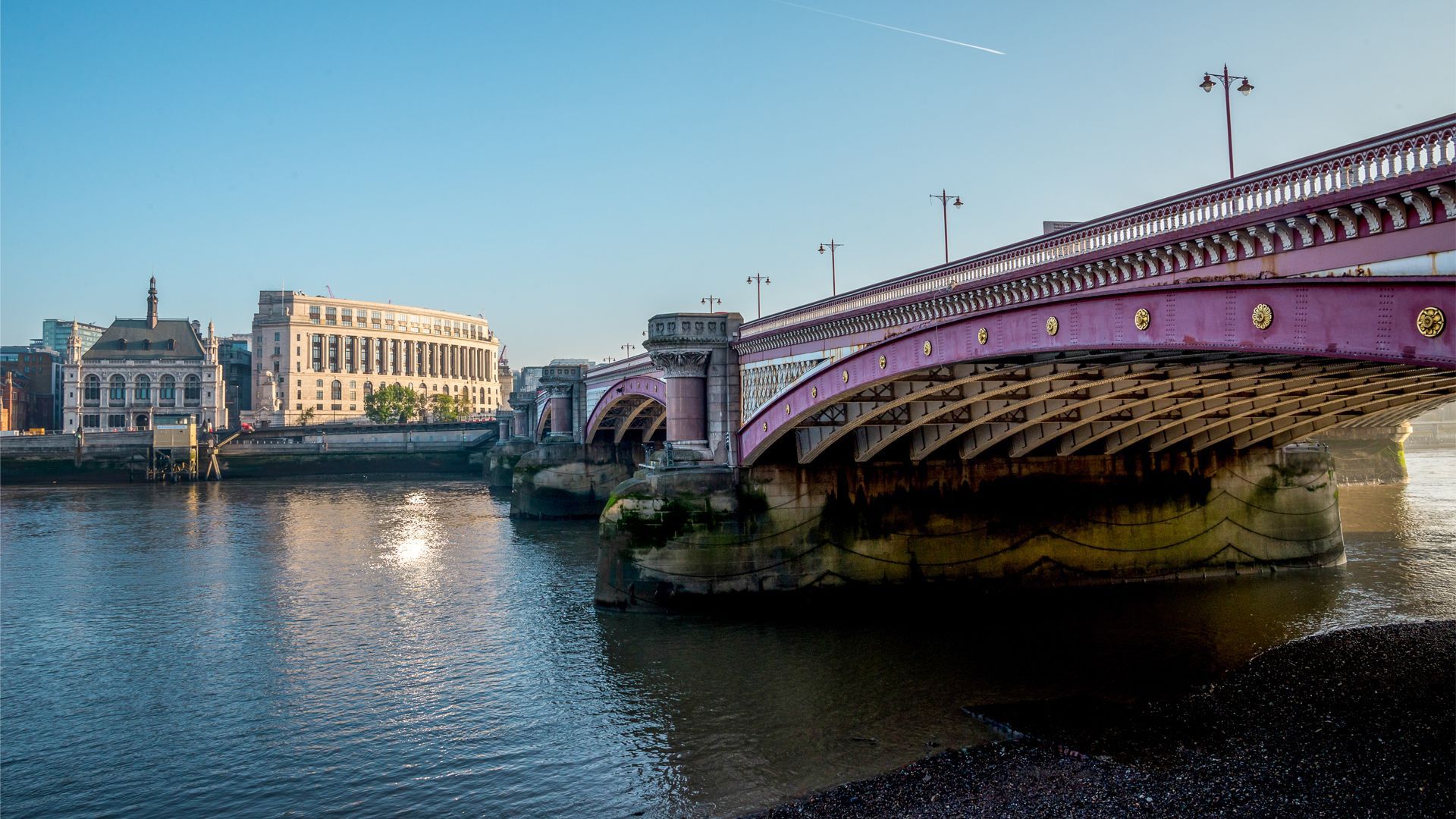 View of Blackfriars Bridge spanning the River Thames with the prominent 100 Victoria Embankment building in the background. The building features a classic architectural style with large windows and a light stone facade, illuminated by bright morning sunlight under a clear blue sky.