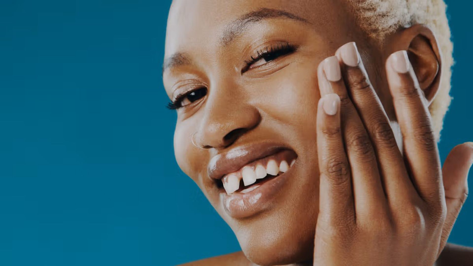 A person with short, light-colored hair and smiling, running their two middle fingers across their face.