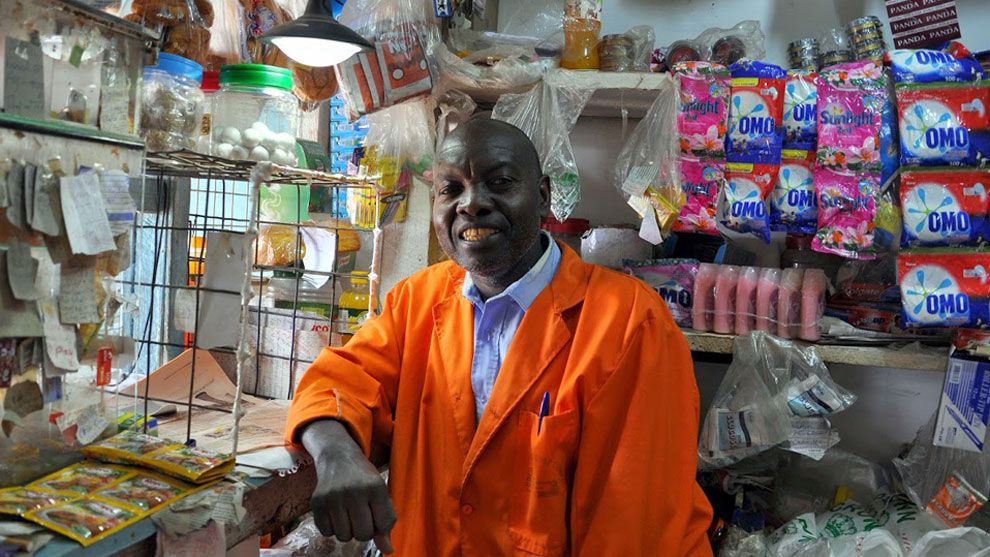A smiling shopkeeper wearing an orange coat stands inside a small convenience store. Behind him, shelves are stocked with various products, including ‘OMO’ and ‘Sunlight’.