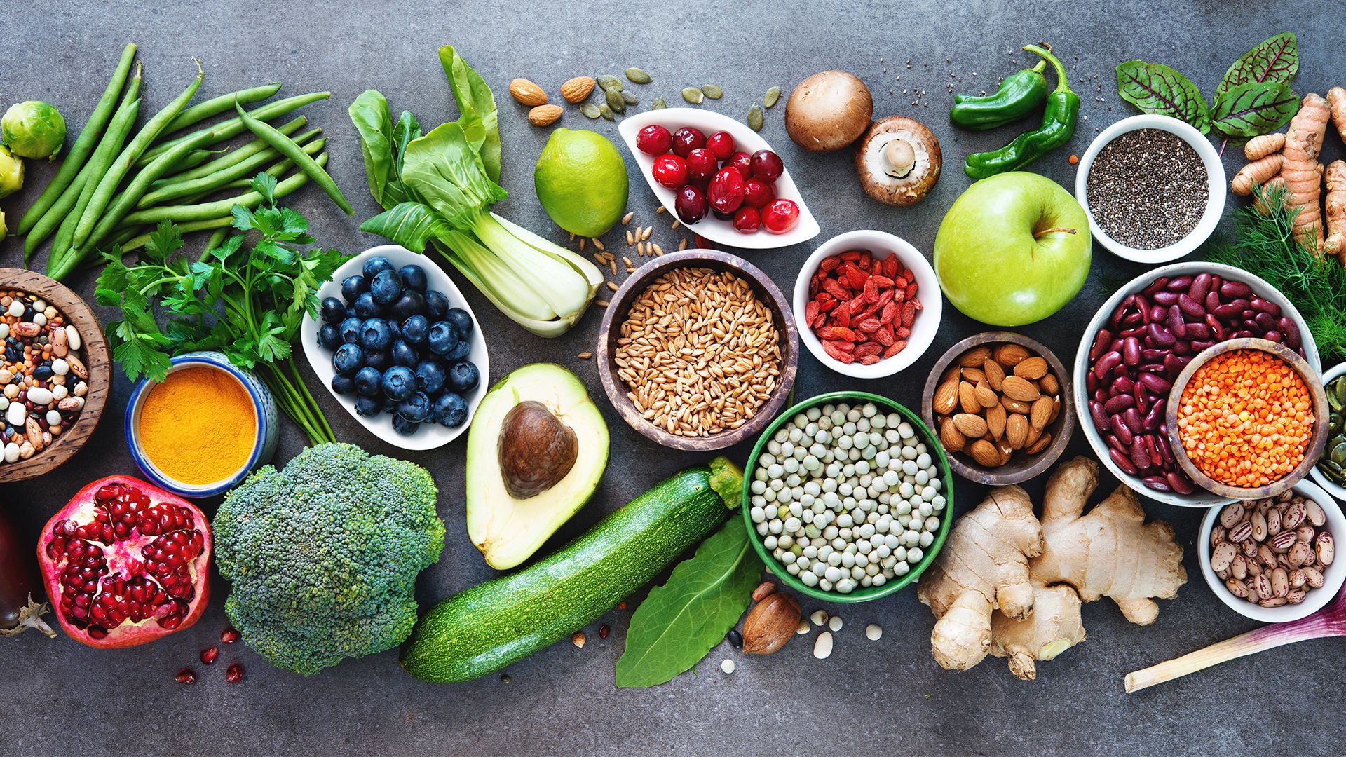 An assortment of fresh fruits, vegetables, legumes, grains, seeds, and spices arranged on a grey surface. Visible foods include green beans, parsley, blueberries, pomegranate, broccoli, avocado, courgette, apple, ginger, turmeric, mushrooms, tomatoes, lentils, kidney beans, almonds, chia seeds, and more, displayed in bowls and loose on the table.