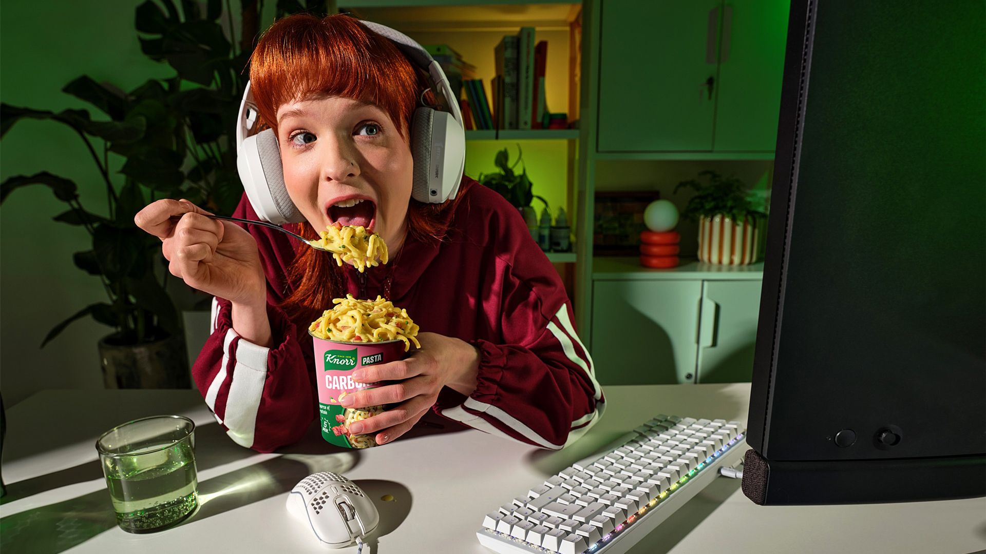 A person wearing large white headphones is eating instant noodles from a cup labelled 'Knorr' while sitting at a desk. The desk has a glass of water, a white computer keyboard, and a mouse and we can see the back of a computer monitor. The background includes shelves with books and plants, and the scene is lit with green ambient lighting.