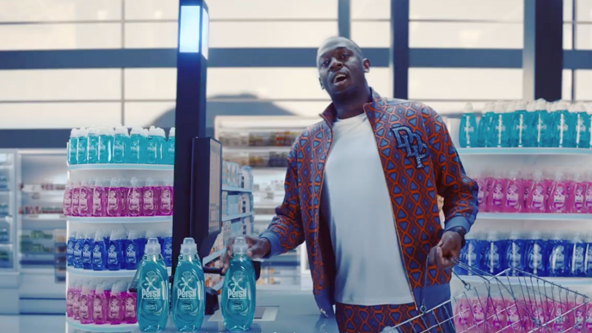 Usain Bolt standing at a checkout counter in a brightly lit store, wearing a patterned jacket and white shirt, holding a bottle of Persil detergent. Shelves in the background are stocked with colourful bottles of cleaning products in pink, blue, and turquoise.