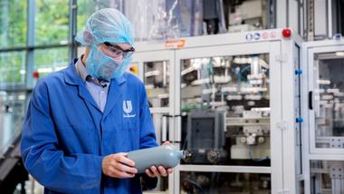A Unilever packaging expert wearing protective clothing and glasses in an industrial setting, inspecting a plastic bottle in front of manufacturing or packaging machinery. The Unilever logo is visible on the person’s coat.