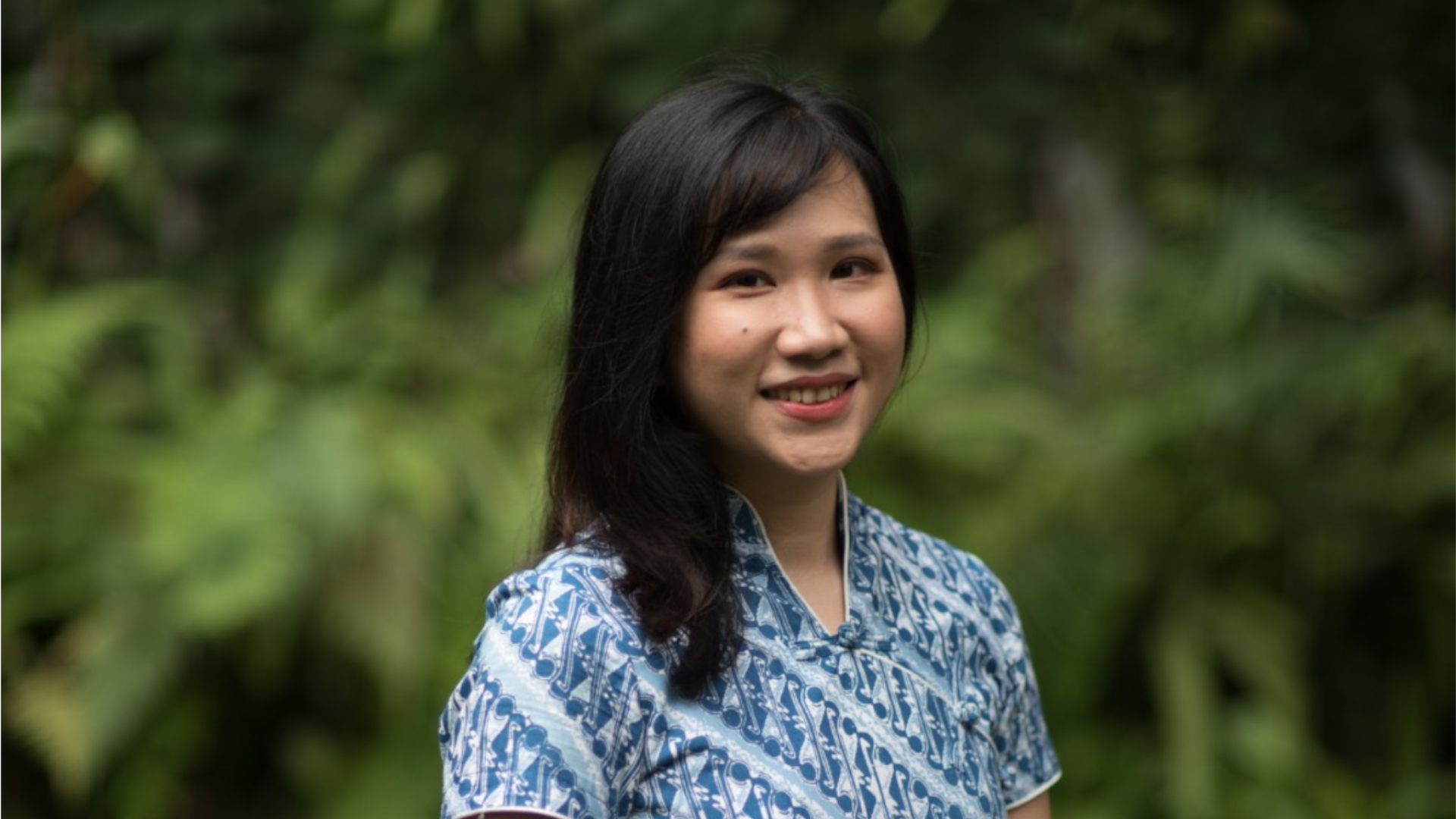 Picture of Lana Kristanto. She has dark hair and is wearing a blue patterned dress against a forest backdrop.