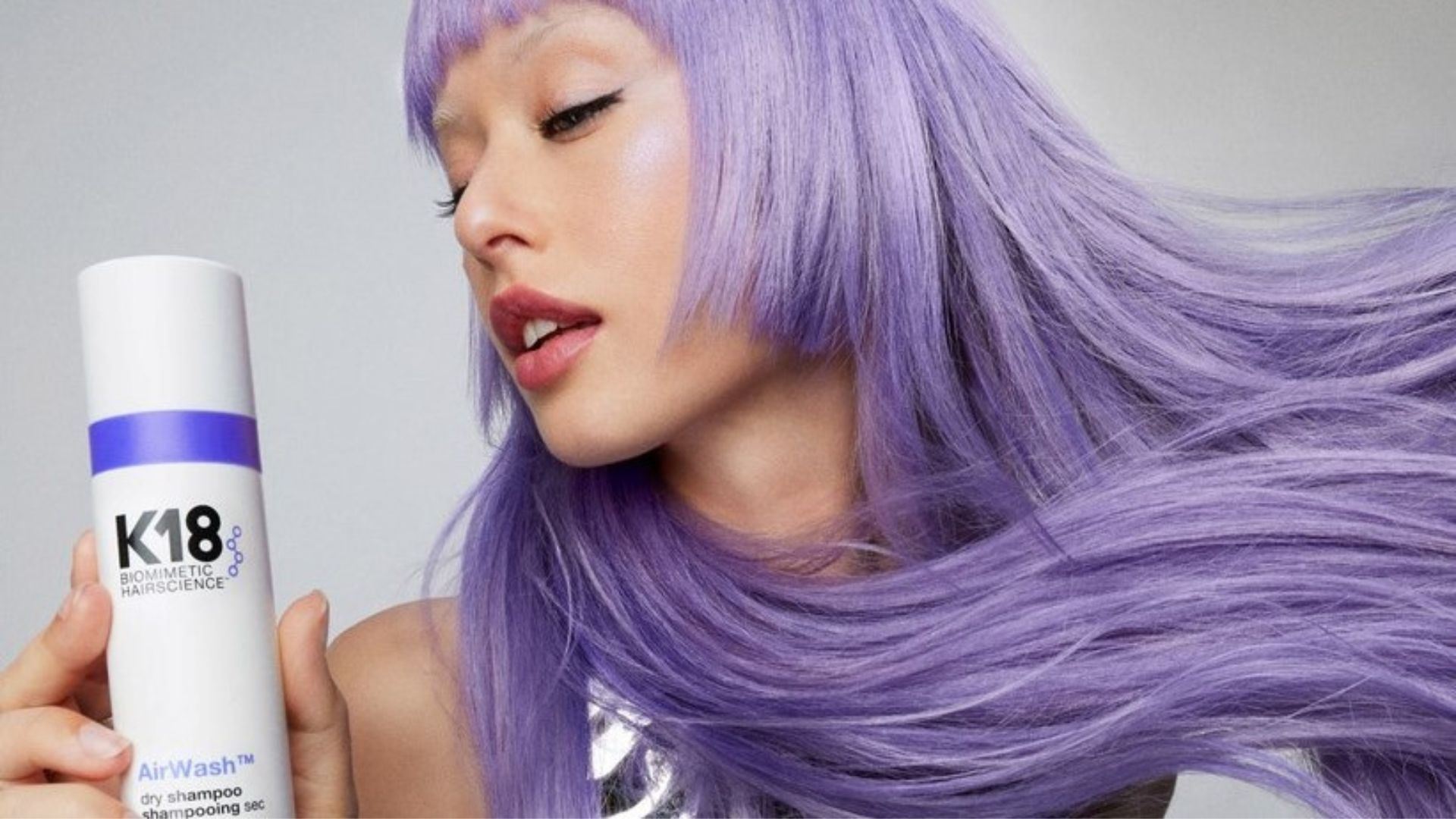 A woman with long violet hair and winged eyeliner holding a bottle of K18 Airwash Dry Shampoo.