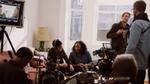 A film crew is taking a break in a light-filled living room with camera equipment on a low table and a screen on a tripod.